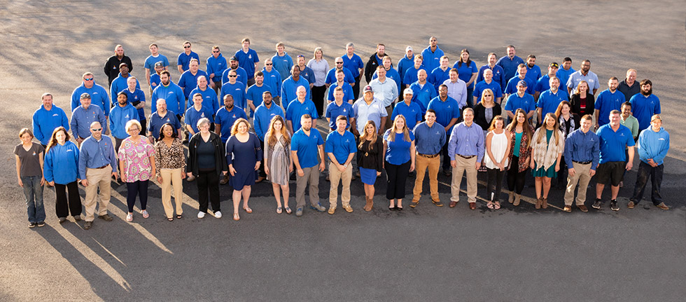 Aerial photo of Raleigh Heating & Air staff standing together in a parking lot, most wearing bright blue shirts.